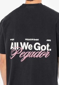 Tricou negru din bumbac cu text imprimat alb și roz pe spate: "Tot ce avem. Pegador" împreună cu detalii ale logo-ului deasupra.
