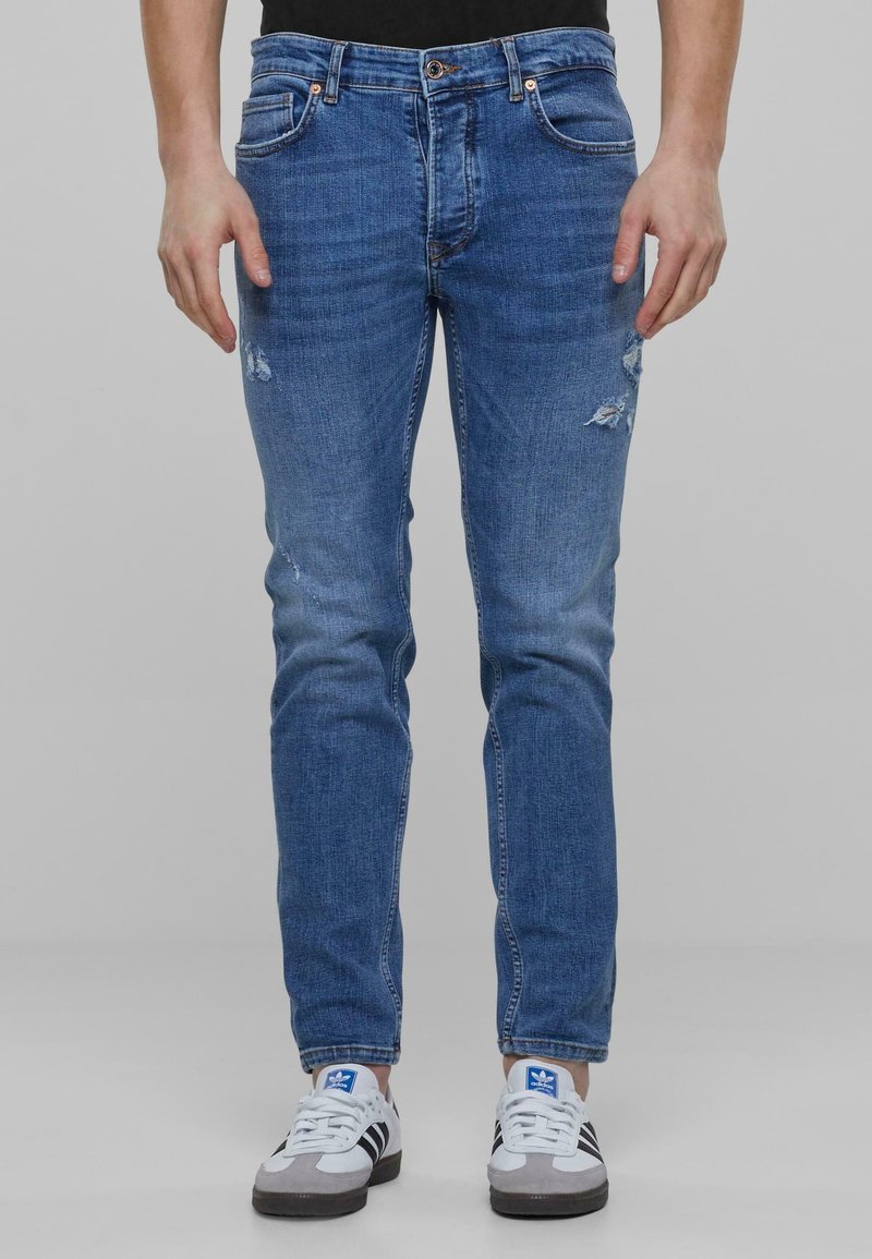 Jean en denim bleu, coupe ajustée. Caractérisé par des déchirures aux cuisses, cinq poches, rivets en cuivre et une texture douce. Style décontracté.