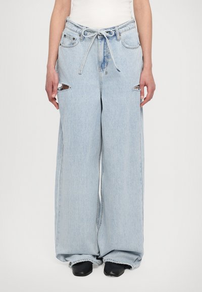 Jean large en denim bleu clair avec une taille haute, cordon de serrage et accents déchirés sur le devant. Finition à bords bruts au niveau des poignets.