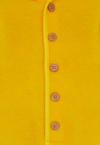 Tessuto giallo con una morbida texture, caratterizzato da sei bottoni marroni sulla parte anteriore e bordo giallo a contrasto lungo la cucitura.
