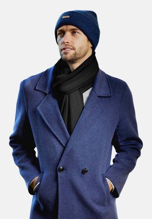 Beanie a costine blu, sciarpa nera a costine e cappotto blu doppiopetto con rever. Il cappotto presenta chiusura con bottoni e tasche laterali.