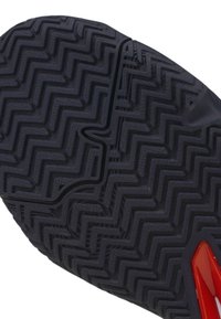 Suela de zapato de goma negra que presenta un patrón de banda de rodadura en zigzag y un acento rojo en el talón, con un surco central para flexibilidad.