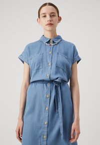 En ljusblå denimklänning med krage, korta ärmar, två bröstfickor och knytmidja, med träknappar.