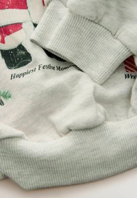 Gaiši pelēks sporta krekls ar ribotām aprocēm, kam ir izdrukāts svētku dizains. Mīksta tekstūra, nedaudz redzami krokojumi un uzraksts "Vislaimīgākie svētku mirkļi."
