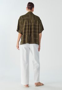 Camicia oversize khaki con motivo geometrico bianco, maniche corte e yoke posteriore. Abbinata a pantaloni larghi bianchi e sandali piatti.