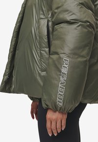Olijfgroene pufferjack met een glanzende afwerking, oversized pasvorm en zwarte ritszakken. "PEGADOR" is verticaal in het wit gedrukt op de mouw.
