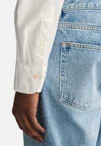 Poignet de chemise à manches longues blanche avec des détails de bouton, associé à un jean en denim bleu clair avec des coutures contrastées et une poche arrière.