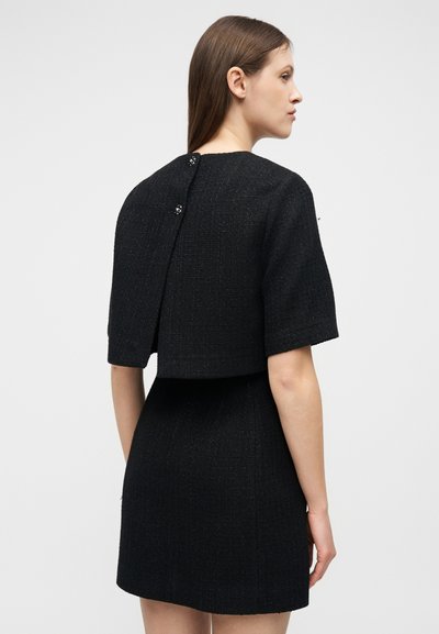 Γυναίκα με ίσια καστανά μαλλιά φοράει ένα μαύρο κοντομάνικο cropped σακάκι με υφή πάνω από μια ταιριαστή κοντή φούστα, με την πλάτη προς τα εμπρός.
