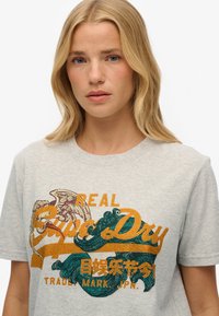 Grå bomuld T-shirt med farverigt tryk med gyldne og grønne accenter, teksten "REAL" og bølger, korte ærmer og rund hals.