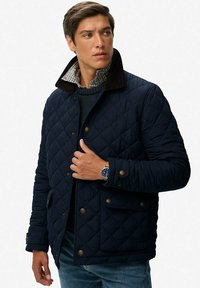 Chaqueta acolchada azul marino con un collar de pana marrón, botones en la parte delantera y bolsillos laterales. Lleva encima un suéter azul marino y una camisa de cuadros con cuello.