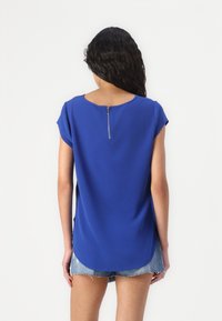 Haut bleu à manches courtes avec un col rond et un détail zippé dans le dos, fabriqué en tissu lisse. Porté avec un short en denim.