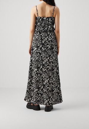 Vrouw die een zwart-witte, abstracte, mouwloze maxi-jurk met dunne bandjes draagt en zwarte, chunky sandalen, staande met de rug naar de camera gericht.