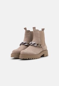 Botas de tobillo de ante beige con suela gruesa, que presentan un detalle decorativo de cadena en la parte delantera y paneles laterales elásticos para facilitar su uso.