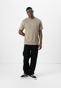 Short-sleeve, light brown cotton t-shirt with logo on chest, paired with black loose-fitting pants and black sneakers with white accents.