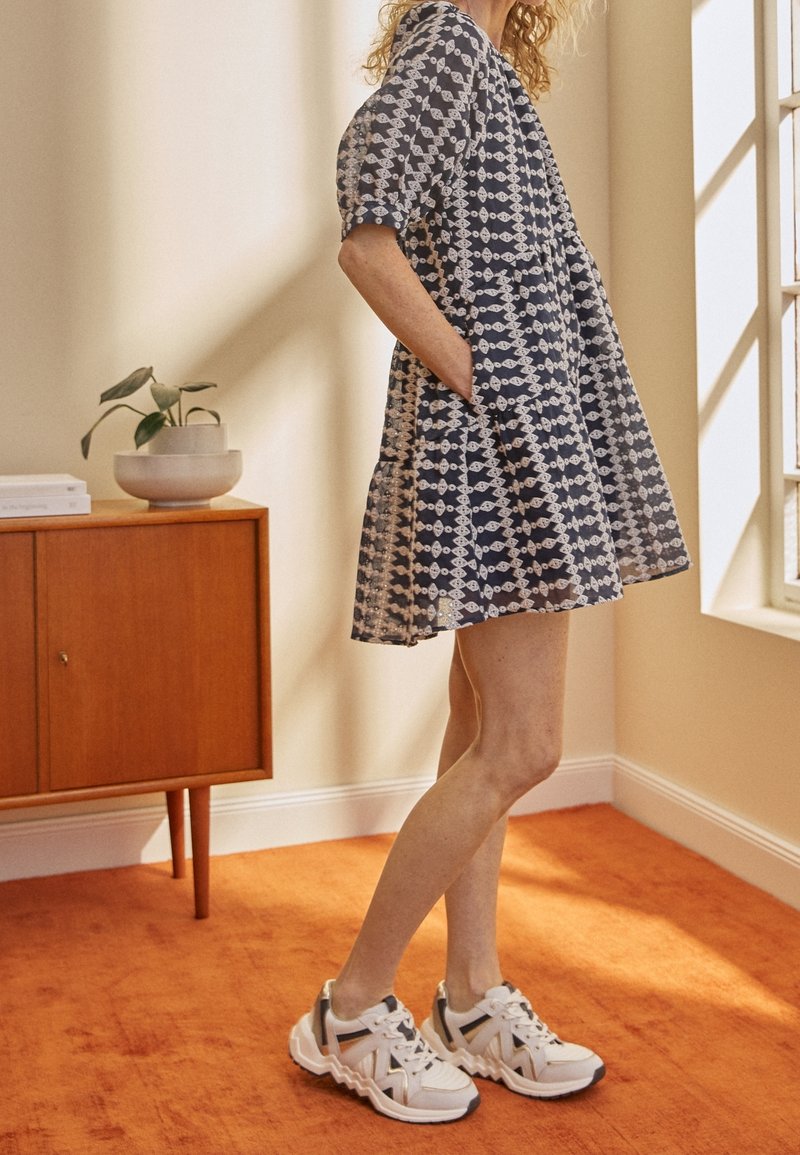 Femme en robe à motifs longueur genou et baskets blanches debout à l'intérieur près d'une armoire en bois et d'une fenêtre sur un tapis orange.