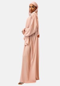 Abaya en satin rose avec des manches longues et un drapé sur le côté. Un hijab assorti complète le look, avec une texture lisse et des détails de design minimaux.