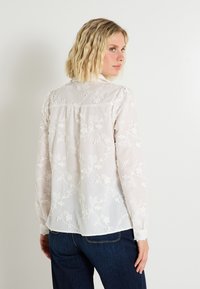 Blouse blanche et transparente avec broderies florales, manches longues, poignets à boutons et un dos plissé. Associée à un jean en denim foncé.