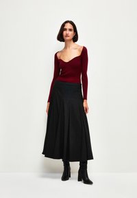 Haut à manches longues côtelé bordeaux avec un décolleté cœur, associé à une jupe plissée noire. La mannequin porte des bottes noires jusqu'aux genoux.