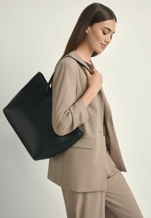 Kobieta w beżowym garniturze niosąca dużą czarną skórzaną torbę na ramieniu, patrząca w dół z lekkim uśmiechem na tle jednolitego tła.
