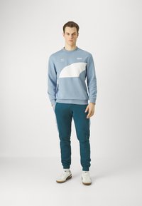 Felpa azzurro chiaro con accenti curvi bianchi e logo, abbinata a pantaloni jogger turchesi con polsini elasticizzati e sneakers bianche.