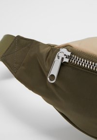 Gros plan d'une fermeture éclair argentée sur un sac en tissu kaki vert et beige avec des détails cousus sur la sangle.