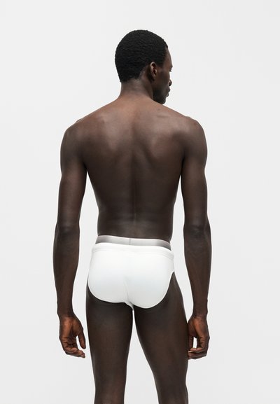 Άνδρας με σκούρο δέρμα, γυρισμένος από την πλάτη, φοράει λευκά εσώρουχα, στέκεται μπροστά σε απλό ανοιχτό γκρι φόντο.
