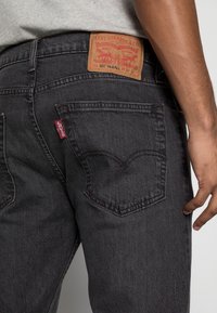 Close-up van een persoon die zwarte Levi's jeans draagt, met de nadruk op de achterzak en het zichtbare Levi's logo-tag en leren merklabel op de taille.