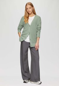 Hellgrüner gerippter Cardigan mit langen Ärmeln und Knopfverschluss, getragen über einem weißen T-Shirt und grauen weit geschnittenen Hosen.