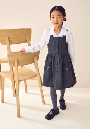Fille portant une robe tablier grise, un chemisier blanc, des collants gris et des chaussures noires, debout avec la main posée sur une chaise en bois sur un fond beige.