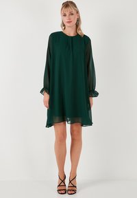 Tamnozelena chiffon haljina s dugim, prozirnim rukavima i slobodnim krojem. Ima okrugli izrez i nabore na manžetama.