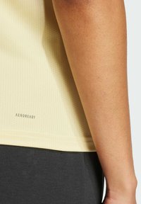 Gelbes, perforiertes Sportshirt aus leichtem Material, mit "AEROREADY"-Logo am unteren Rand, kombiniert mit dunkelgrauen Hosen.