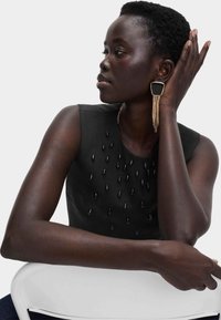 Top negro sin mangas con adornos en forma de gota de agua brillantes, combinado con un gran pendiente rectangular de oro y negro. Textura suave.