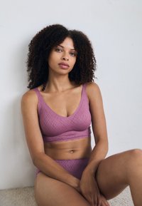 Lavender bra and brief set made of textured fabric; features a scalloped hem and a V-neck design. Model seated against a neutral background.