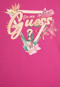 Růžové tričko s lesklým logem "Guess", květinovými grafikami s růžovými květinami a tropickými listy a geometrickým emblémem.