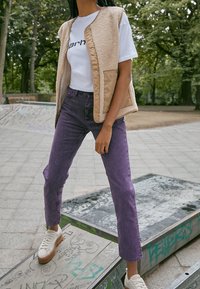 Beige väliosa, jossa on sherpa-vuori, valkoisen t-paidan päällä, jossa on mustaa tekstiä. Violettiset suoralahkeiset farkut ja valkoiset lenkkarit, joissa on ruskea pohja. Ulkoilmamiljöö.