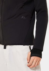 Black puffer jacket with textured sleeve, featuring a fitted design, ribbed accents, and discreet branding on the chest. Zippered pocket visible.