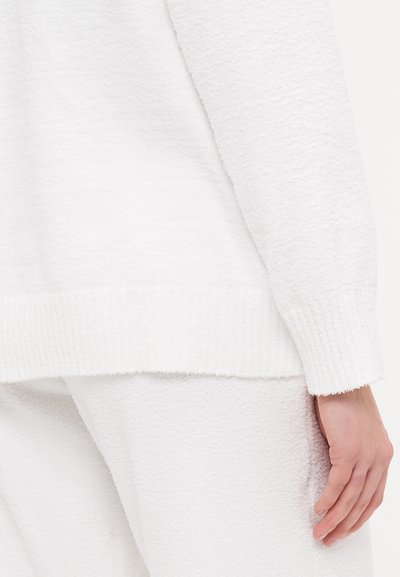 White textured sweater with ribbed cuffs, featuring a soft, fluffy material. Close-up shows hand resting on the garment's surface.