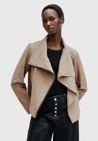 Giacca in suede marrone con colletto drappeggiato, chiusura con zip e tasche laterali, abbinata a pantaloni neri a vita alta con dettagli a bottone.