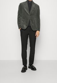 Ciemnozielona marynarka ze sztruksu z dwoma kieszeniami, zestawiona z prążkowanym czarnym golfem, czarnymi spodniami i czarnymi mokasynami z złotym akcentem.