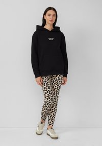 Zwarte hoodie met een voorvak en witte tekst, gecombineerd met crèmekleurige en zwarte luipaardprint leggings en beige sneakers.