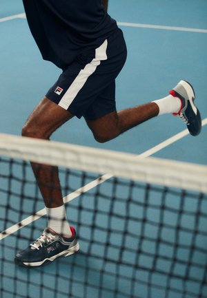 Blå idrettsshorts med hvite sidestriper, kombinert med en marineblå og rød sneakers, som løper på en blå tennisbane, foten i midt-steg.