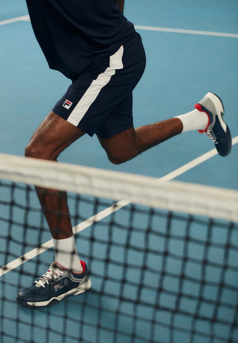 Sinised spordilühikesed püksid valgete külgtriipudega, koos tumesiniste ja punaste tossudega, joostes sinisel tenniseväljakul, jalg keskel sammu.