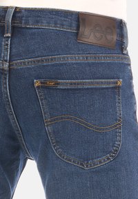 Donkerblauwe denim jeans met een kenmerkend bruin leren label met de tekst "Lee." Versterkte stiksels en traditionele achterzakken.