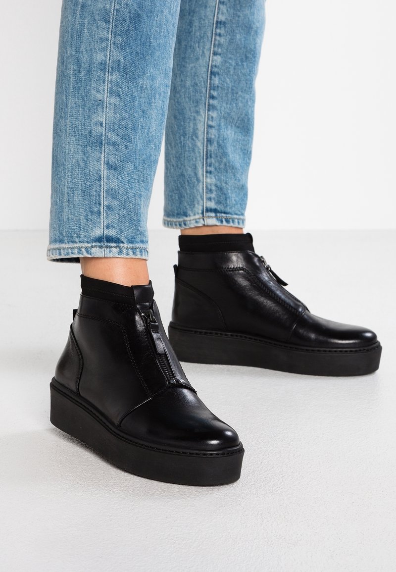 Bottines à plateforme en cuir noir avec fermeture éclair à l'avant, portées avec un jean bleu clair, présentées sur un sol blanc.