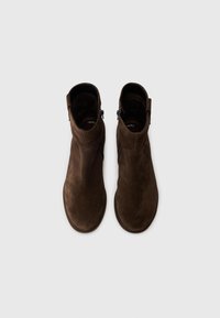 Hnědé semišové kotníkové boty s elegantním designem, vybavené bočním zipem a minimálními švy, umístěné na ploché podrážce.