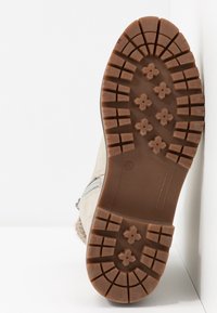 Semelle d'une botte beige avec une bande de roulement en caoutchouc brun comprenant des crampons carrés et en forme de fleurs, présentée sur un fond blanc.