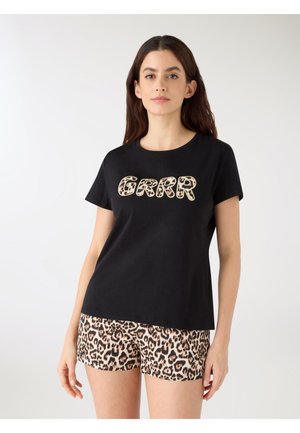 Kobieta w czarnej koszulce z napisem "GRRR" w panterkowy wzór i dopasowanych szortach w panterkę, stojąca na białym tle.