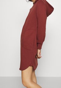 Czerwona sukienka z kapturem o luźnym kroju, z kieszeniami typu kangur, długimi rękawami i lekko zaokrąglonym dołem po bokach. Miękka tekstura materiału.