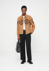 Giacca di lana marrone con motivi a diamante, camicia beige, pantaloni neri, con una borsa in pelle nera, abbinata a scarpe nere.