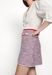 Femme portant une blouse blanche avec un col en dentelle et des manches bouffantes, associée à une jupe à carreaux rouge, blanc et bleu marine avec une poche avant.